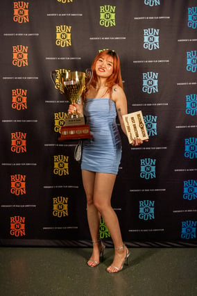 A young Asian woman holds a big trophy in front of a film festival backdrop that reads 'Run N Gun'