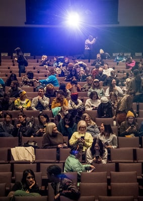 A movie theatre room, with the audience getting to their seats and the projector shining a flare