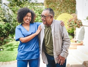 A nurse stands beside an elderly man outside, both smiling and engaged in conversation in a sunny ou