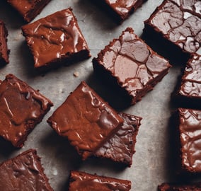 A stack of rich, chocolatey brownies is placed on a slate board, with chunks of dark chocolate scattered around. The brownies have a dense, moist texture, and the background includes a blurred view of a kitchen setting.