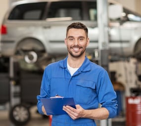 Homem de macacão azul com uma prancheta dentro de uma Oficina Mecânica com um carro ao fundo