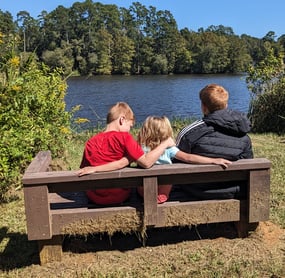 3 kids sitting on a bench in front of a lake