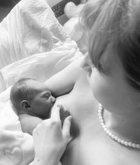 Black and white top view of new mom nursing baby in bed. Baby holds mother's finger as she gazes down with love.