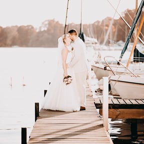 Hochzeitspaar steht auf einem Steg am Wasser, umgeben von Booten und Natur.