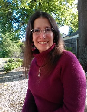 brunette woman wearing a purplish-pink sweater and glasses, outdoors, looking at camera and smiling