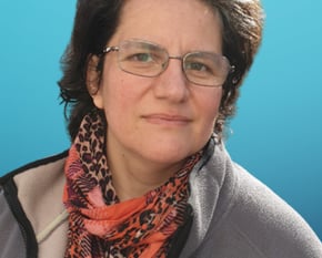 A woman with short dark hair and glasses looks toward the camera. She wears a neutral-toned jacket.