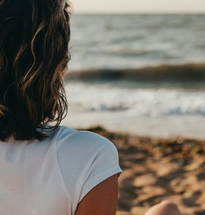 Femme seule vue de dos, assise sur la plage, face à la mer. Un enfant joue au loin, au bord de l'eau