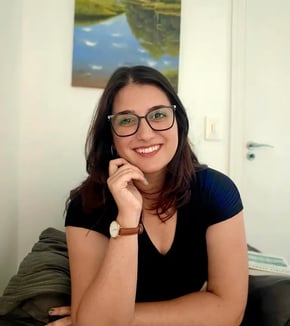 Psicóloga Rafaela Antoniazzi sorrindo, vestindo óculos e blusa preta, apoiando o rosto na mão. Seção Sobre.