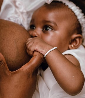 Bébé fille vêtue de blanc noire tétant le sein de sa maman et lui tenant le doigt