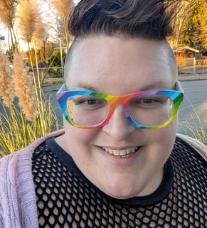 A knitter with short brown hair and rainbow glasses smiling.