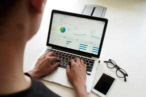 Homem analisando gráficos em um laptop, com óculos e smartphone sobre a mesa.