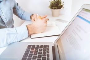 Homem escrevendo em um caderno, com laptop e celular sobre a mesa.