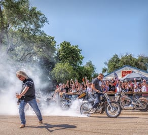a man playing a guitar in front of a crowd with a man burning out on his motorcycle.