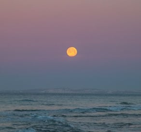 a full moon rising over the ocean with a pink sky