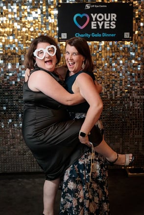 Two ladies striking a pose in front of the SweetAz Co. silver shimmer wall at the Layton Vision Foundation Charity Gala in Br