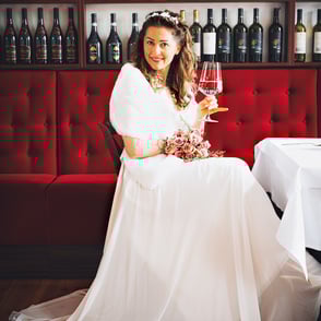 Frau in weißem Kleid mit rosafarbenem Blumenstrauß und Rotwein, vor einer Bar.