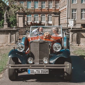 Ein retro Auto mit zwei Personen sitzt davor, lächelnd in der Sonne.