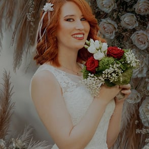 Braut mit rotem Haar hält einen Blumenstrauß, lächelt vor einer dekorativen Kulisse.