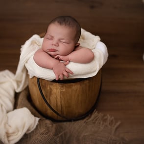 Photo de bébé endormi, délicatement posé dans un sceau en bois, entouré d’un wrap clair