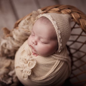 Photo de bébé endormi, emmailloté dans un wrap avec des noeuds et posé dans un panier en osier tout en douceur