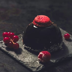 A glossy chocolate entremet cake topped with a red macaron, accompanied by red currants on a dark slate surface.