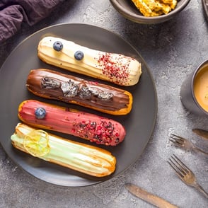 Four colorful eclairs with different toppings on a dark plate.