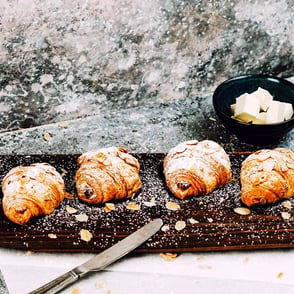 Four chocolate croissants on a wooden board with almond flakes and powdered sugar sprinkled on top.