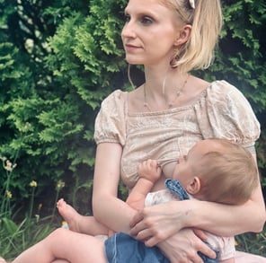 Mother sitting in front of a green bush, wearing a bow in her hair, holding and nursing her baby & looking away peacefully