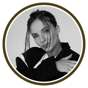 A person in a stylish black outfit poses with one arm wrapped around themselves against a muted backdrop.