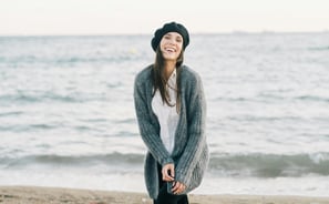 Woman smiling and standing on a beach wearing a sweater and beret.