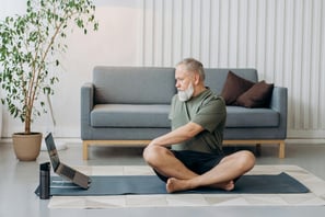 Hombre adulto mayor sentado con las piernas cruzadas sobre un tapete de yoga en su sala.