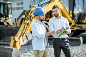 two men in hard hats and hard hats standing in front of a construction site