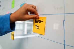 A hand posting a sticky note with the phrase "how-to" written on it.
