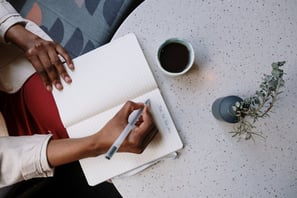 A hand journaling on a notebook that is placed on a table with a cup of tea.
