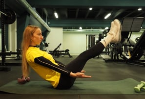 Una donna in maglia gialla esegue l'esercizio 'boat pose' su un tappetino da yoga in palestra, concentrandosi sull'equilibrio