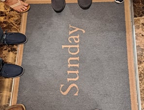 A rug that says "Sunday" with several pairs of feet standing around it