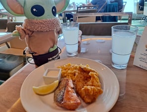 Little green companion beside a plate with salmon and a piece of fried chicken