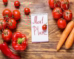 A notepad with "Meal Plan" written on it, surrounded by fresh vegetables on a wooden table.