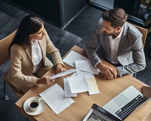 Two business people sitting at a table with papers and laptops