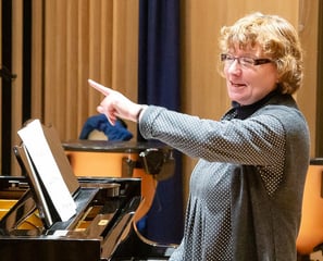 a woman in a gray sweater and glasses is pointing at a piano