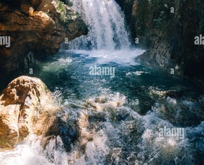 a waterfall in the middle of a river with sun shining through the trees