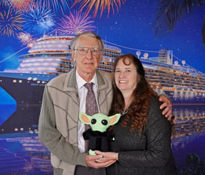 A couple standing in front of a cruise ship backdrop holding a little green companion