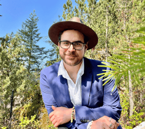 a smiling man in a suit and hat sitting on a bench in a forest