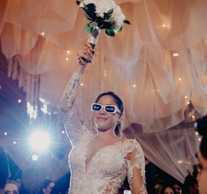 Novia levantando ramo de boda en la mano derecha con lentes de sol con las iniciales de ella y novio