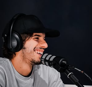 aledima sorride mentre parla al microfono durante un podcast, con cuffie e cappello nero