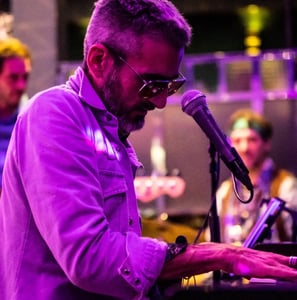 un homme joue du piano et chante lors d'un concert. Il est éclairé d'une lumière violette