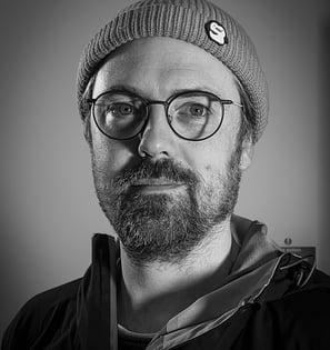 A black and white headshot of a man in his thirties wearing glasses and a hat.