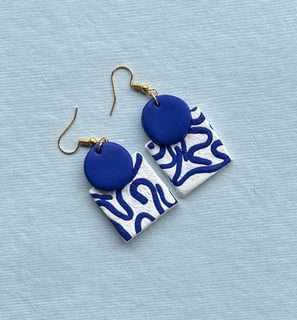 santorini themed pair of white clay earrings with abstract indigo patterns displayed on a sky blue background 