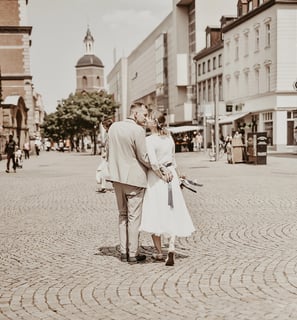Ein Hochzeitsfoto, das ein Paar von hinten beim Küssen zeigt, während sie durch eine gepflasterte Straße gehen. Der Mann träg