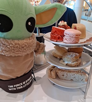 Little Green companion standing beside three tiered plate of mini cakes and tea sandwiches